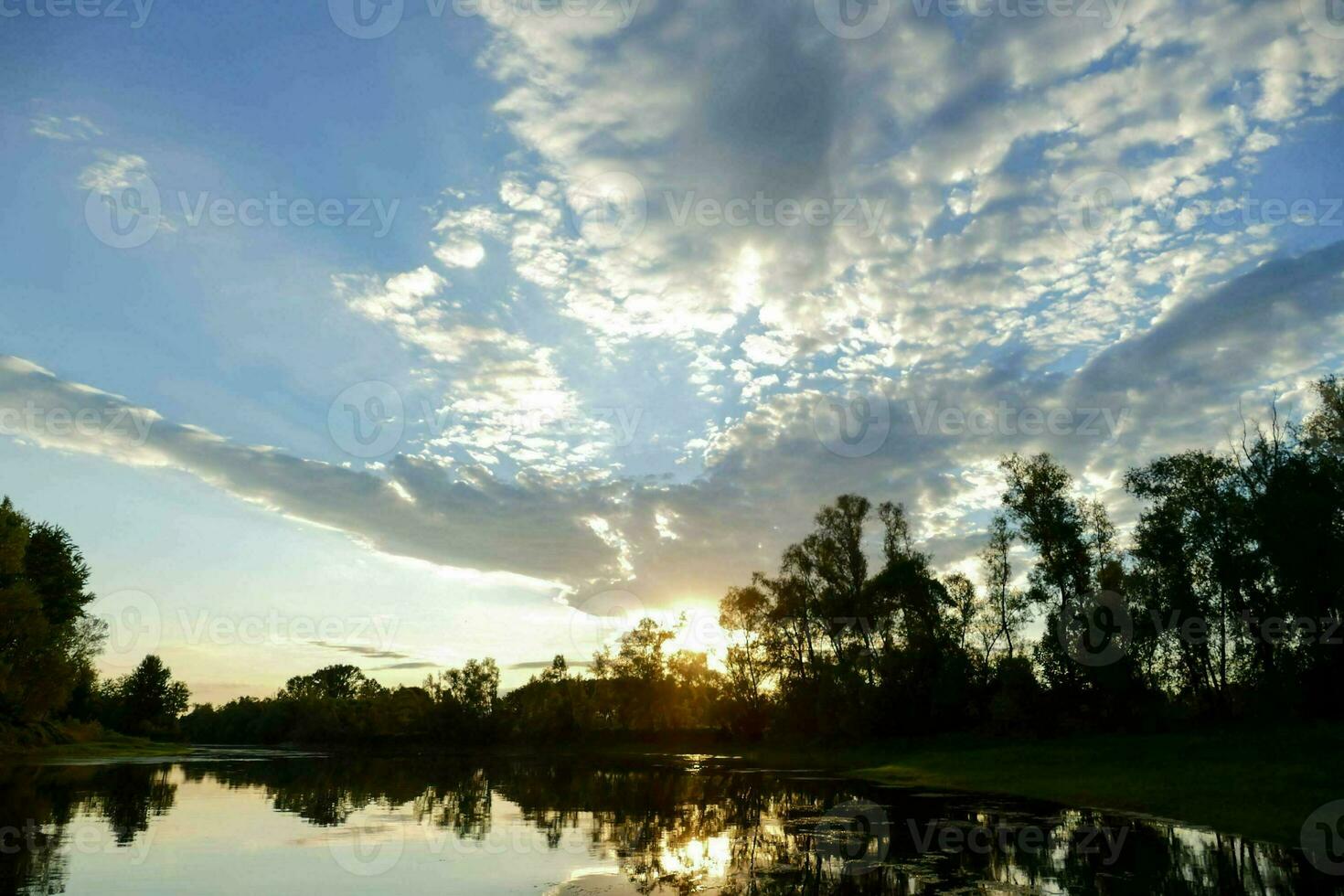 the sun is setting over a lake with trees and clouds photo