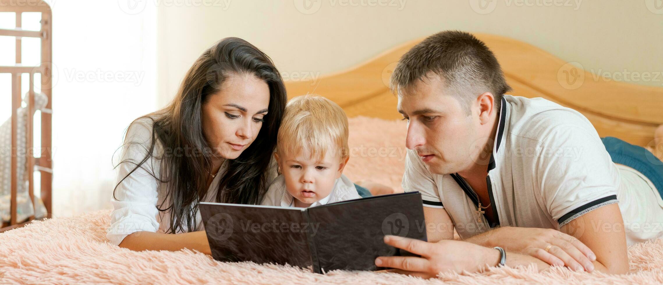 happy family lying on the bed reading a book, mom dad and little son, banner photo