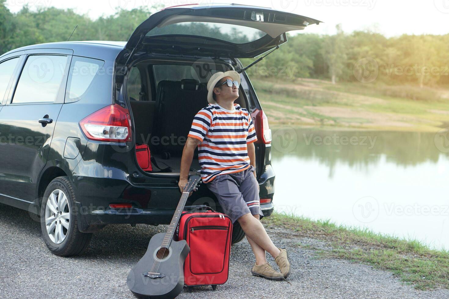 Asian man traveler sit on trunk of car, put guitar and suitcase on the floor, parked car beside the lake.Concept, relax time. Vacation or holiday trip by private vehicle. Give time for yourself. photo