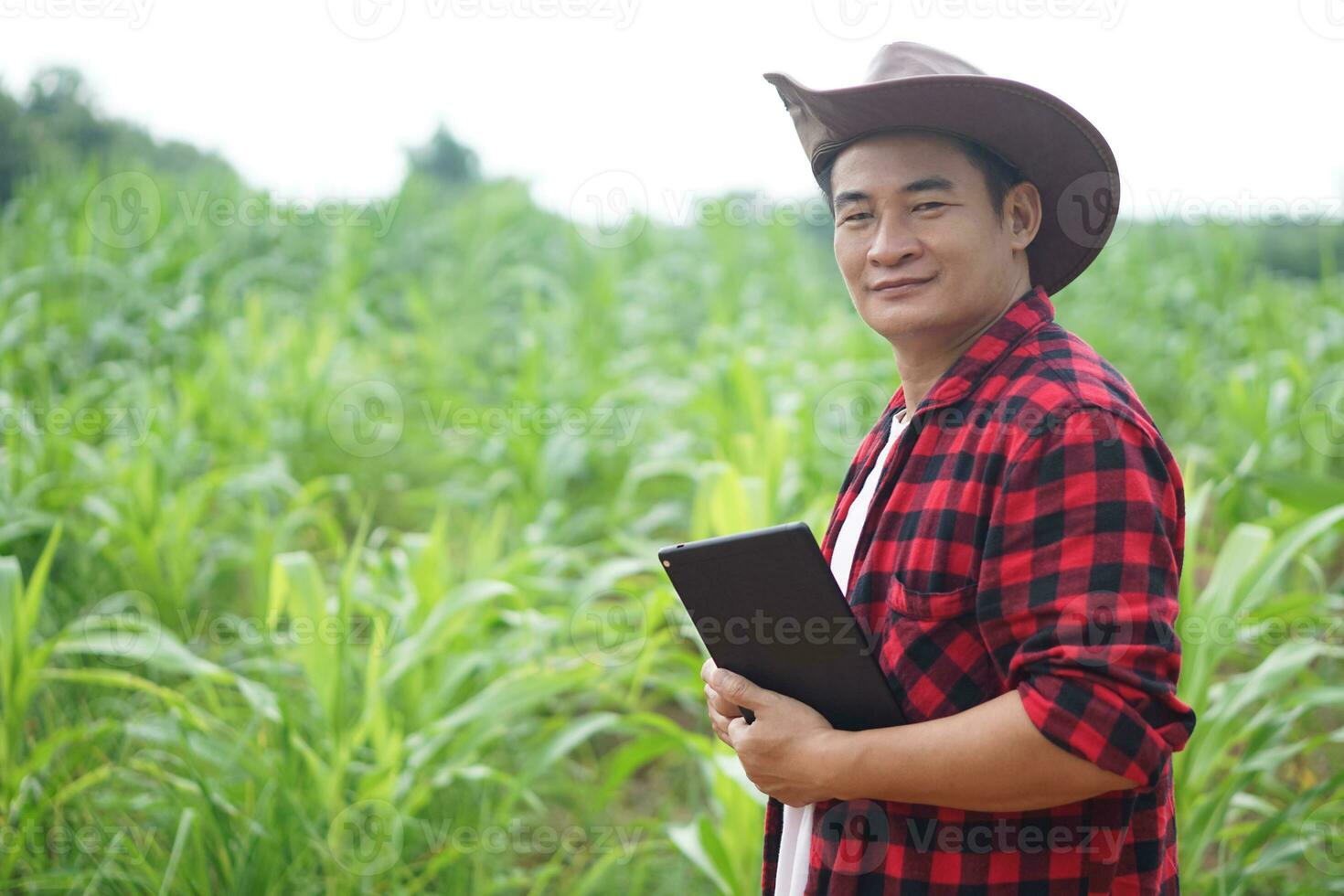 Handsome Asian man farmer is in garden, wears hat, red plaid shirt, holds  smart tablet. Feel confident, proud in crops. Concept, Agriculture occupation, inspect, survey and research. Copy space. photo