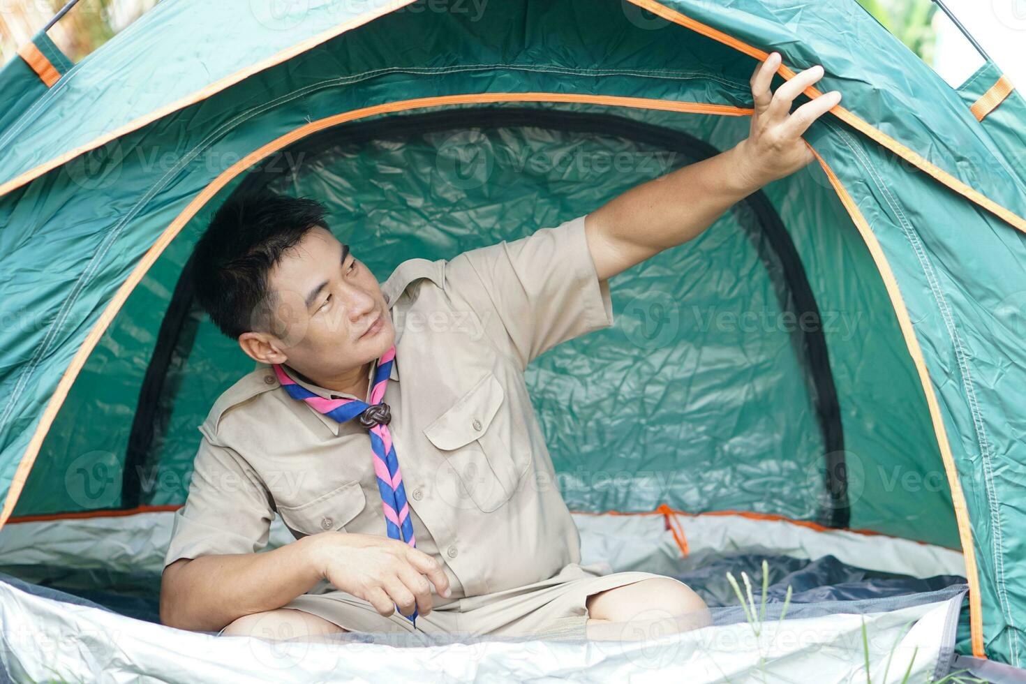 Handsome Asian man wears boy scout uniform sits in camping tent. Concept, accommodation for temporary outdoor sleeping as scout hiking activity. Scout go camping. photo