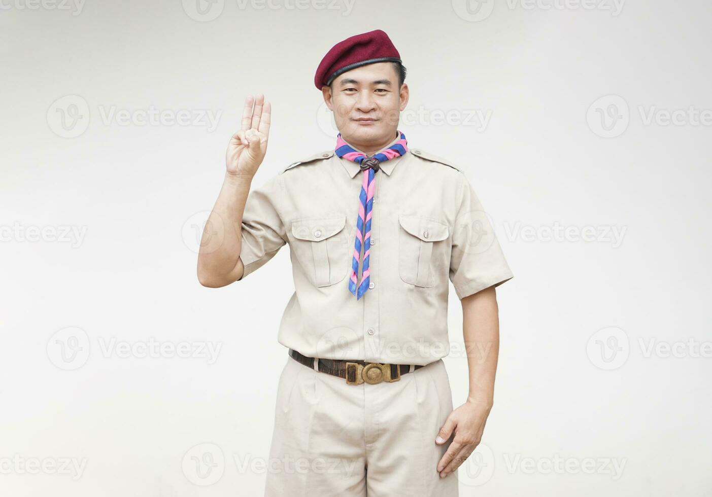 Handsome Asian man wear boy scout uniform, red cap, blue and pink striped scarf, make hand sign symbol of scout for oath. Concept, educational career with uniform in school, Thailand. photo