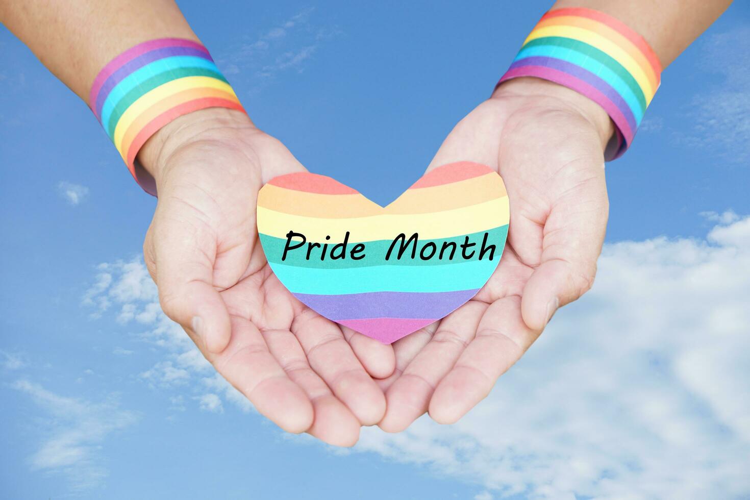 Close up hands wears rainbow color wrist band and holds rainbow heart shape with word Pride Month. Blue sky background. Concept, Pride month for LGBTQ community celebration in June. Human right photo