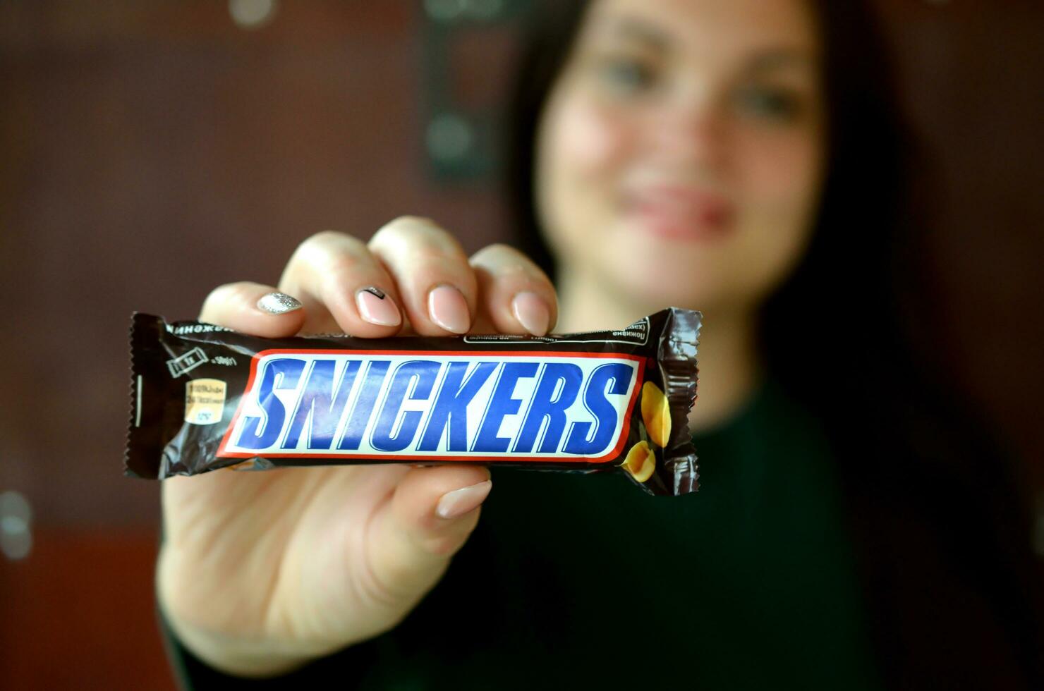 KHARKOV, UKRAINE - OCTOBER 8, 2019 A young caucasian brunette girl shows Snickers chocolate bar in dark room. Snickers chocolate manufactured by Mars photo