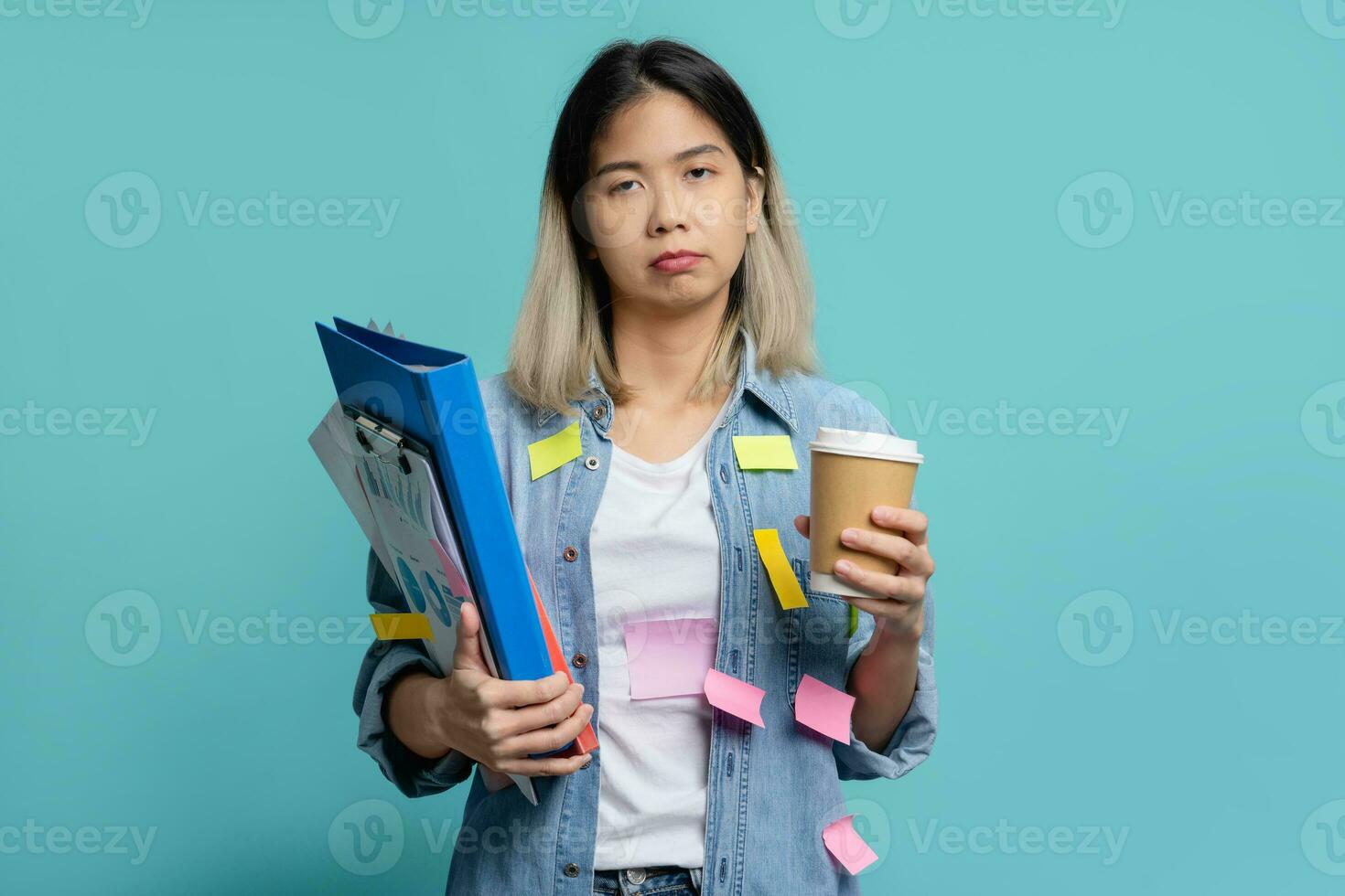 Confused female office worker holding folder and cup of coffee, on her body full of sticky note paper stickers with deadline for completion of research results standing isolated on blue background. photo