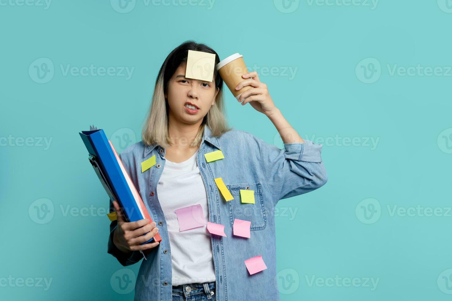 Frustrated Asian female manager holding document file with coffee cup and a sticky note on the face, stand on studio shot blue background. photo