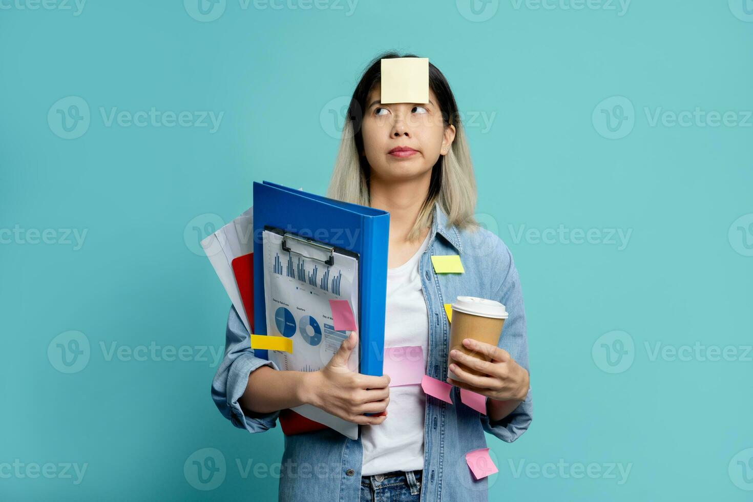 Bored and confused young Asian woman holding document file and notes on her forehead. She stands holding mug of hot coffee and looking sideways isolated on blue background. photo