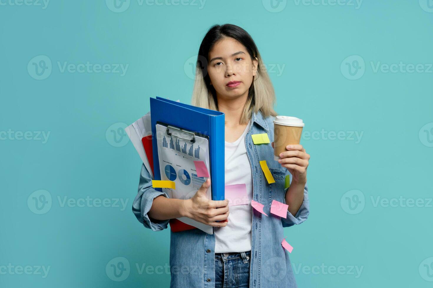 Tired and unhappy asian woman holding many document file has deadline with cup of coffee on blue background in studio. photo