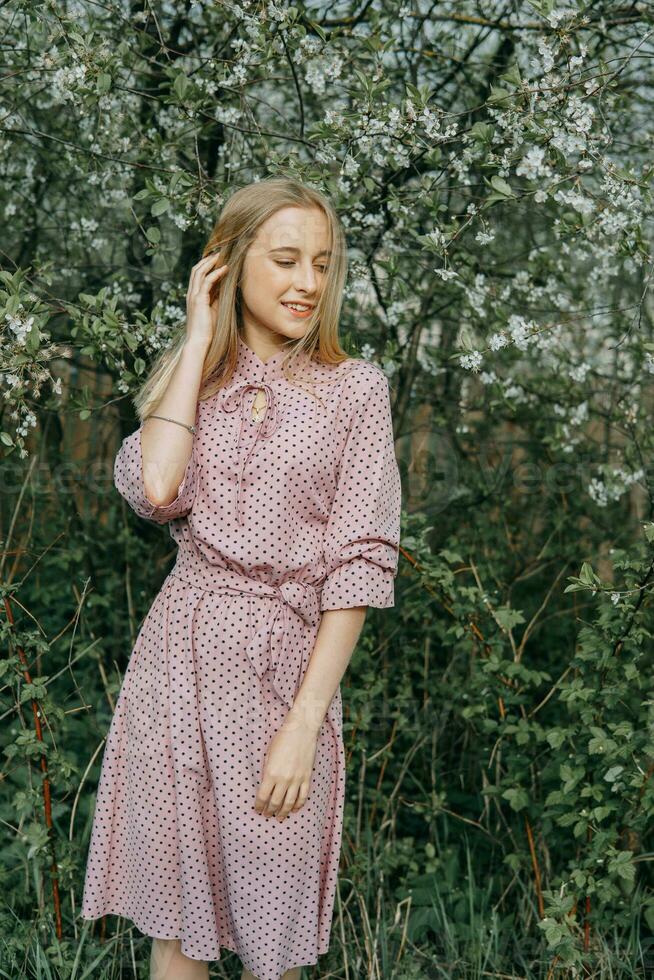 Blonde girl on a spring walk in the garden with cherry blossoms. Female portrait, close-up. A girl in a pink polka dot dress. photo