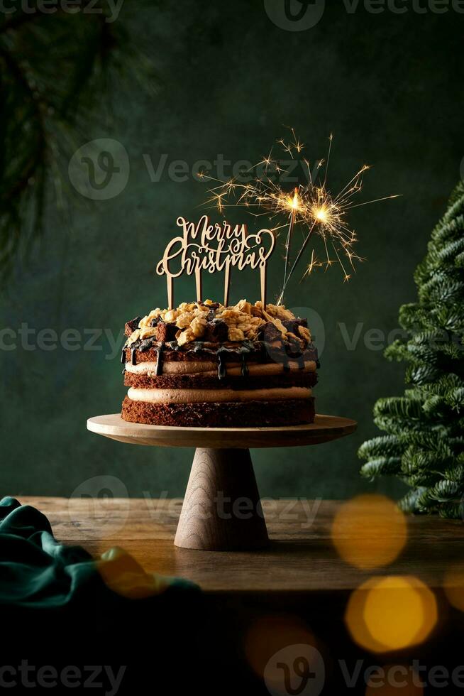 Naked chocolate cake with Merry Christmas decoration and sparklers on a wooden rustic table with green background. Cozy Christmas themed still life. photo