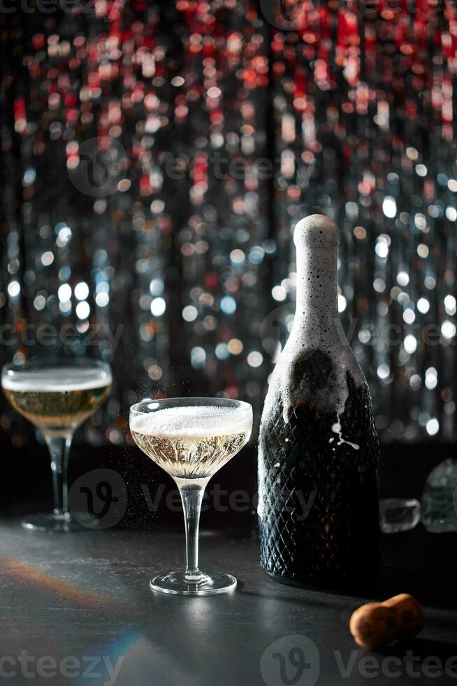 Image of celebration. The champaign bottle splashing next to which are two glasses of champaign. The background of bokeh with colors red and silver. Dark grey stone table. photo