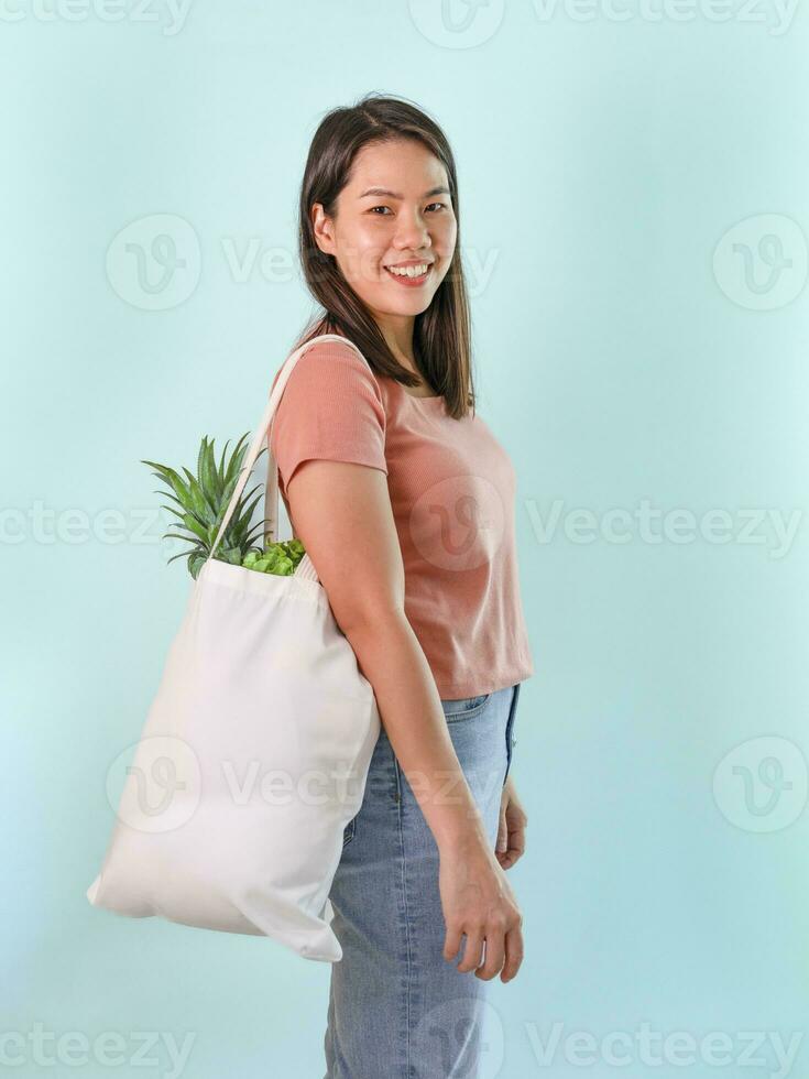 happy young asian housewife carrying reusable cloth bag of fruits and vegetables isolated on blue background,concept for campaign of using reusable bag, reduce waste,stop global warming photo