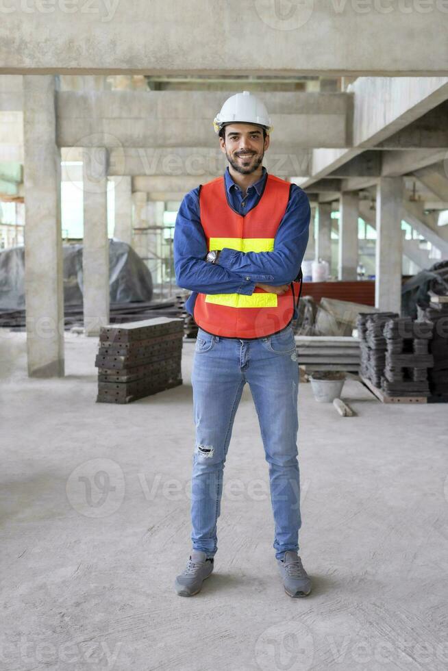 vertical image of attractive young male engineer wearing safety gear,standing arms crossed,smiling confident, background blurred a new building under construction site photo