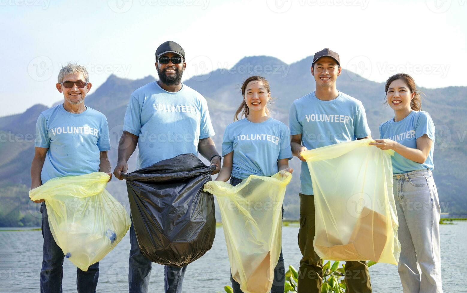 multiethnic people in blue volunteer T-shirt volunteers holding waste sorting plastic bag standing by the river,a diverse group of volunteers join together to cleanup the sand beach together photo
