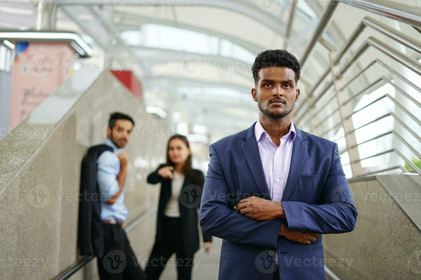 young executive standing with arms crossed is suspiciously, background are employee talking about him as bullying. concept advertising,article,magazine etc for management conflict in the workplace etc photo
