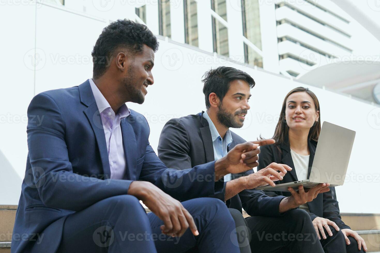 multiracial business colleagues sitting and working outside building, young business people using laptop to discuss work outdoors photo