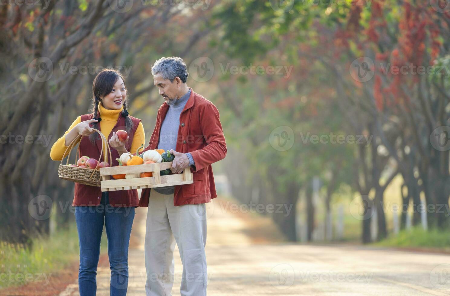 a daughter holding basket of fruits and father carrying wooden crate fill with pumpkins smile and talk about quality of agricultural productivity while walking along the street at farmland in autumn photo