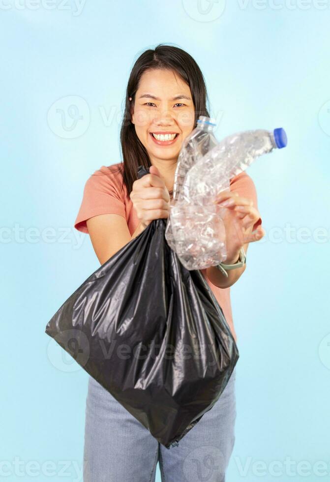 smiling asian housewife one hand holds plastic bag,other hand holds used plastic bottle on blue background,concept of waste management,campaign to separate household waste, support garbage recycling photo