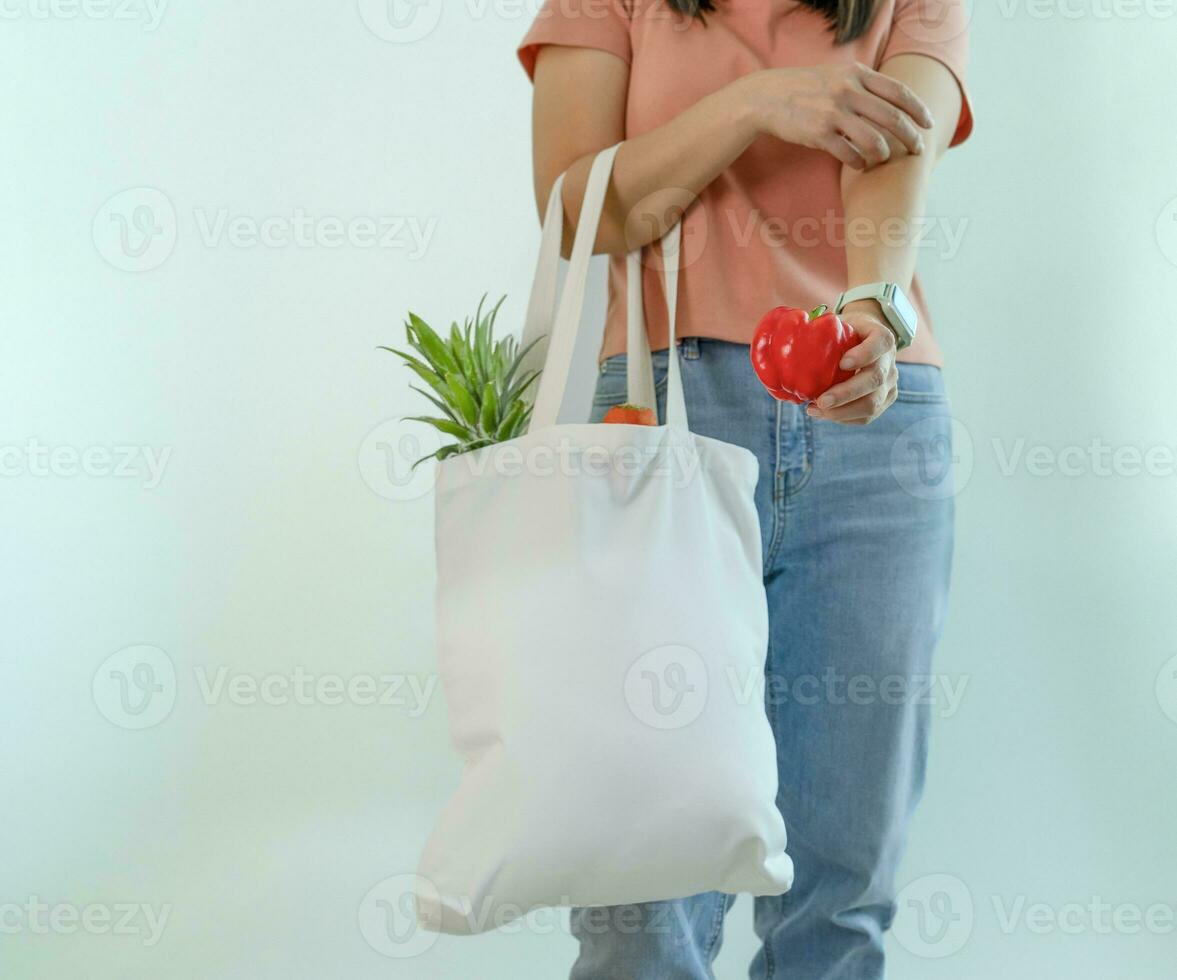 a female one hand holds red bell pepper,other hand carry reusable cloth bag of fruits and vegetables isolated on blue background,concept campaign of using reusable bag,zero waste, stop global warming photo