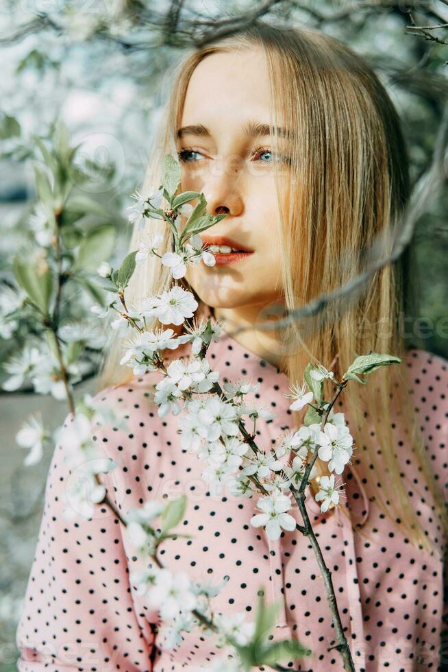 Blonde girl on a spring walk in the garden with cherry blossoms. Female portrait, close-up. A girl in a pink polka dot dress. photo