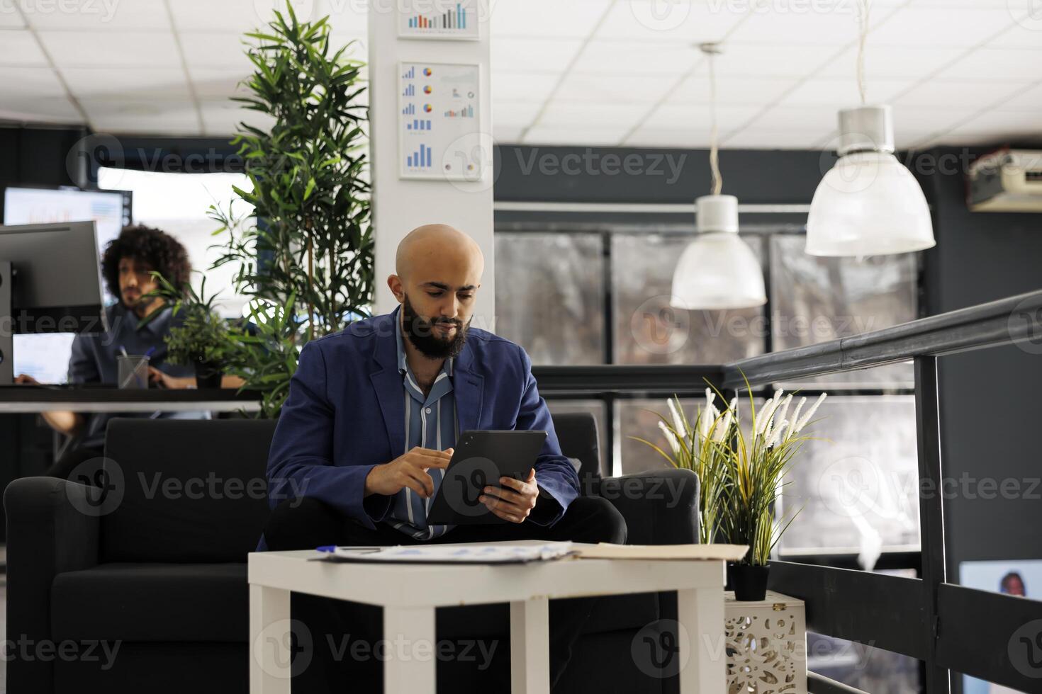 Arab businessman using tablet for digital work in business office. Company man employee scrolling through social media during break while sitting on couch in coworking space photo