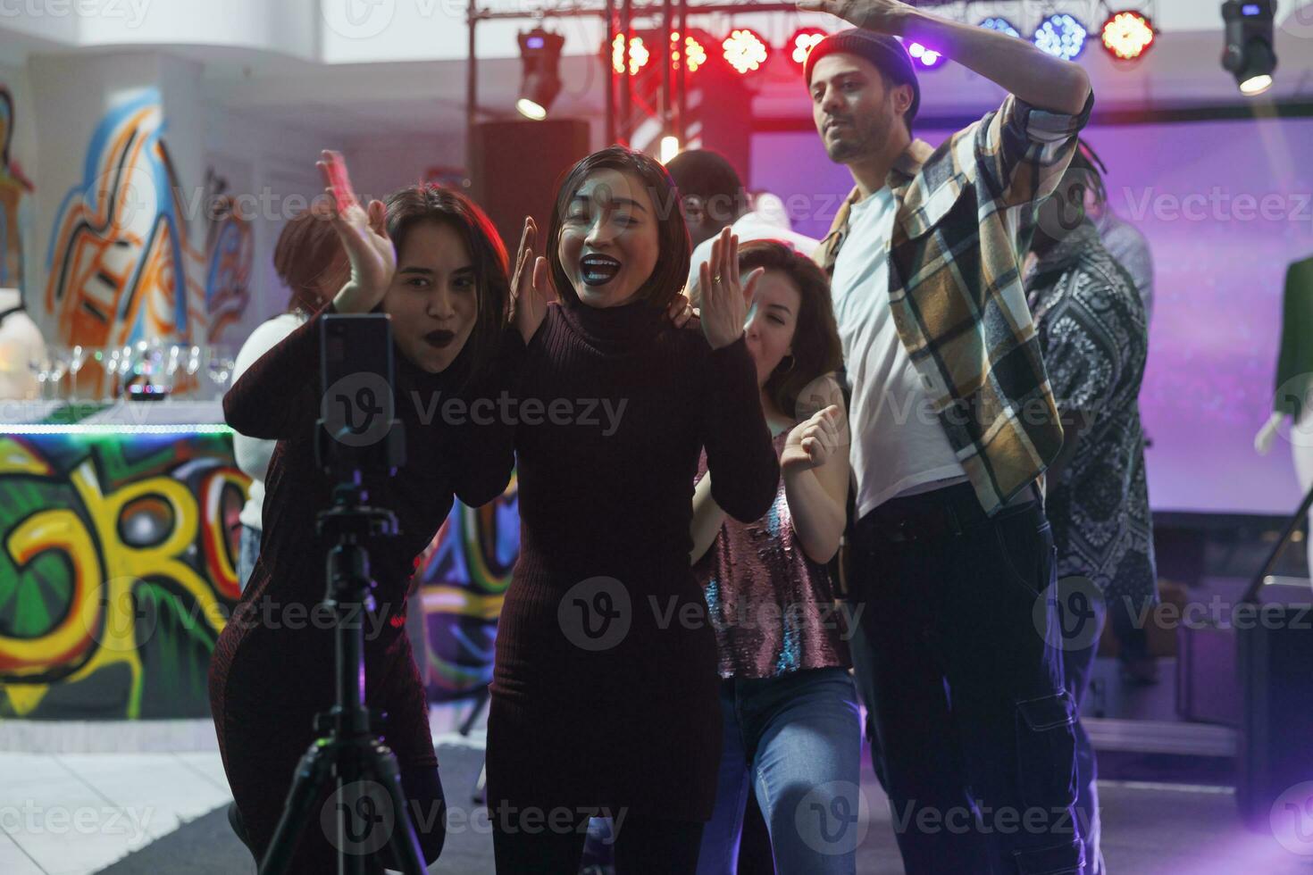 Group of cheerful friends taking photo on smartphone with tripod while partying together in nightclub. Diverse young men and women streaming club discotheque on mobile phone