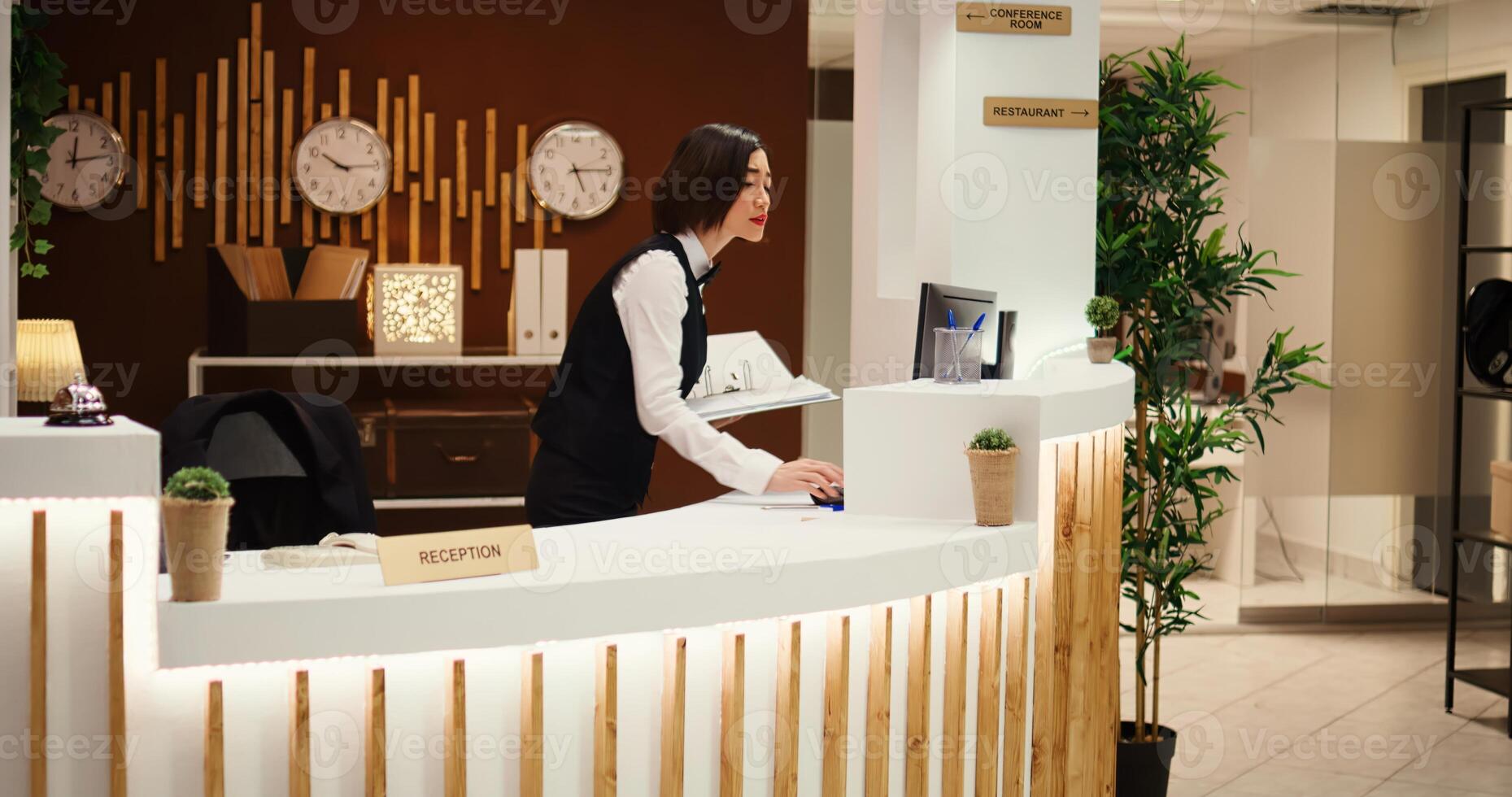 Happy friendly receptionist making phone calls standing at check in counter in welcoming resort lobby. Cheerful professional hotel employee taking booking requests while working at reception desk photo