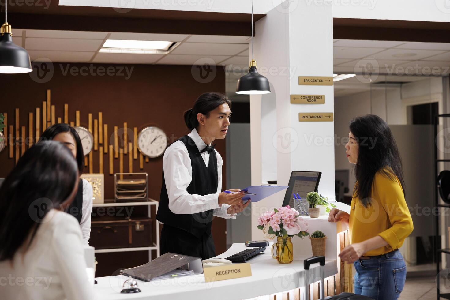 Hotel guest receiving registration form from helpful asian male concierge. Relaxed asian middle aged woman on vacation making necessary preparation for check in at modern resort photo