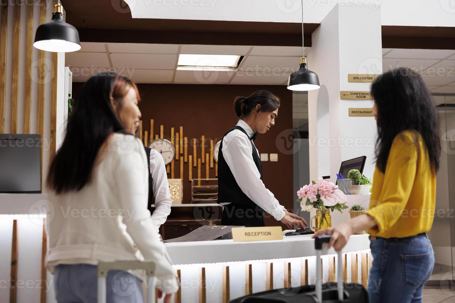Cozy hotel reception with two female guests waiting for concierge to finalize booking procedure. Relaxed customers holding baggages getting ready to check in at lavish resort photo