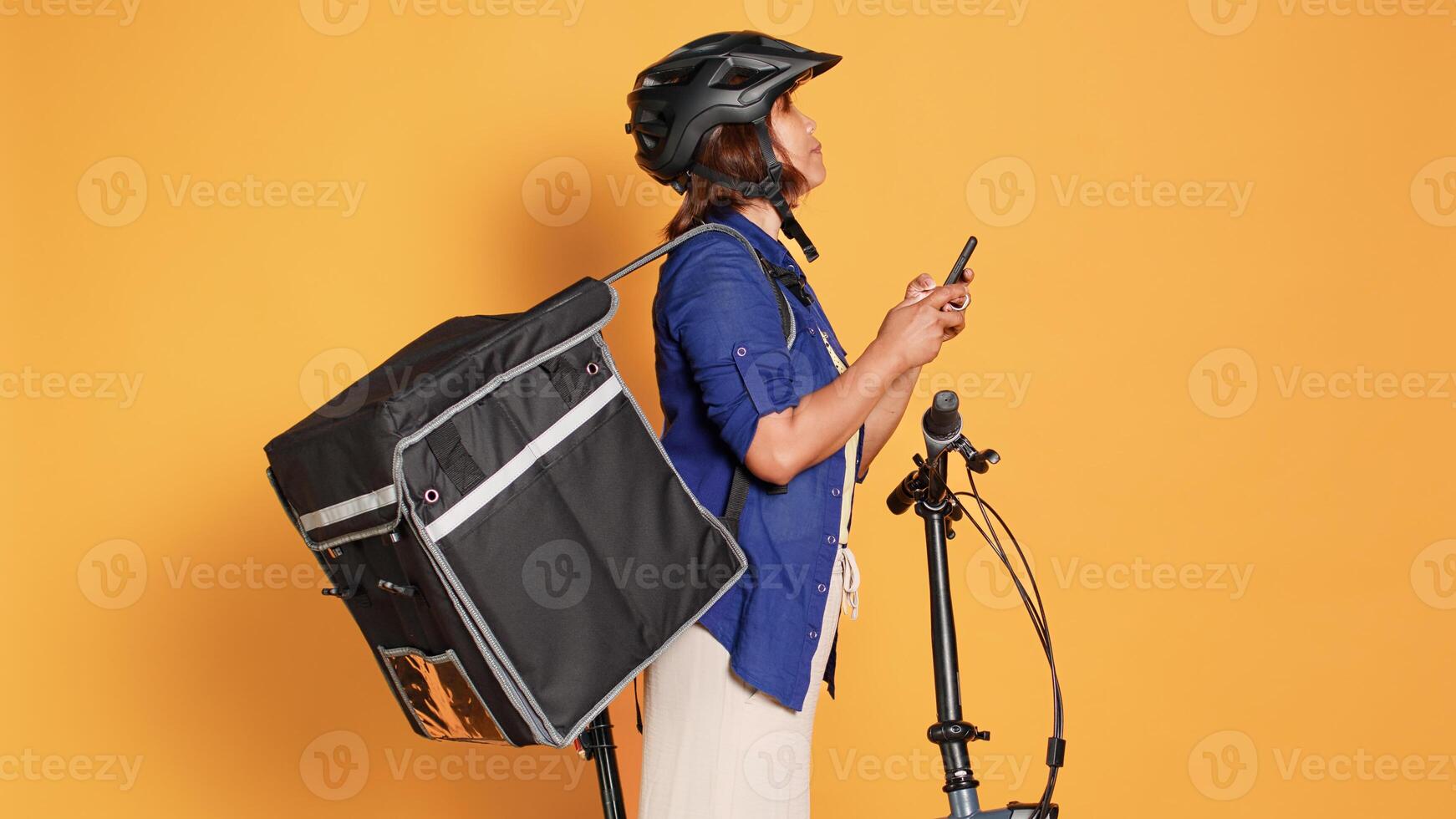 Courier writing down customer order in phone app, preparing to ride bike to client address. Asian cyclist isolated over orange studio background delivering takeaway food using thermal backpack photo