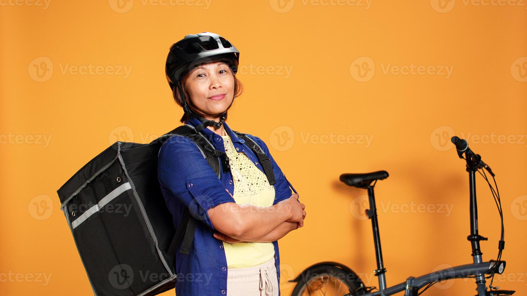 Portrait of courier proud of quick professional finished delivery to customer. Bike rider transporting fast food meal to clients using thermic backpack, isolated over orange studio background photo