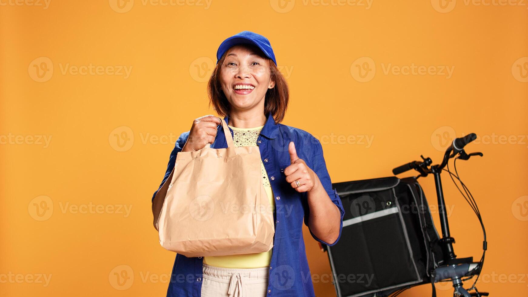 Slow motion portrait shot of bored food delivery bike rider waiting for customer, showing exultant approving gesturing. Asian courier isolated over studio background providing meal order to client photo