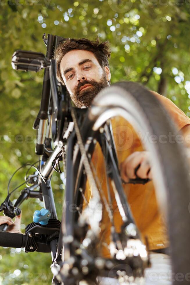 Healthy vibrant caucasian man is seen outdoors doing annual upkeeping and maintenance of modern bicycle. Active young male cyclist checking and ensuring functioning of bike pedal. photo