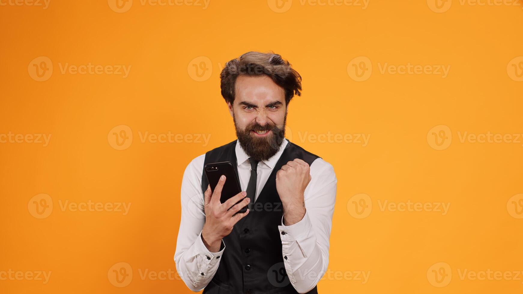 Joyful butler uses smartphone app in studio, feeling happy after reading messages on camera. Confident stylish worker with suit and tie browsing online on mobile phone, working at restaurant. photo