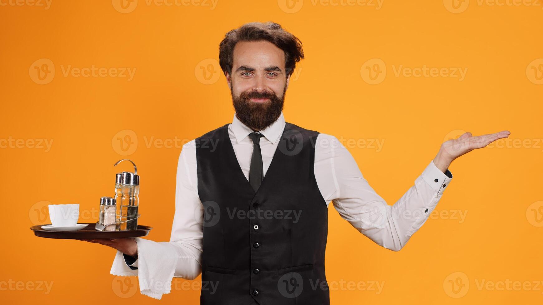 Confident waiter poses with tray in hand, standing against yellow background and working at restaurant. Catering industry worker with metal platter and towel providing outstanding luxury service. photo