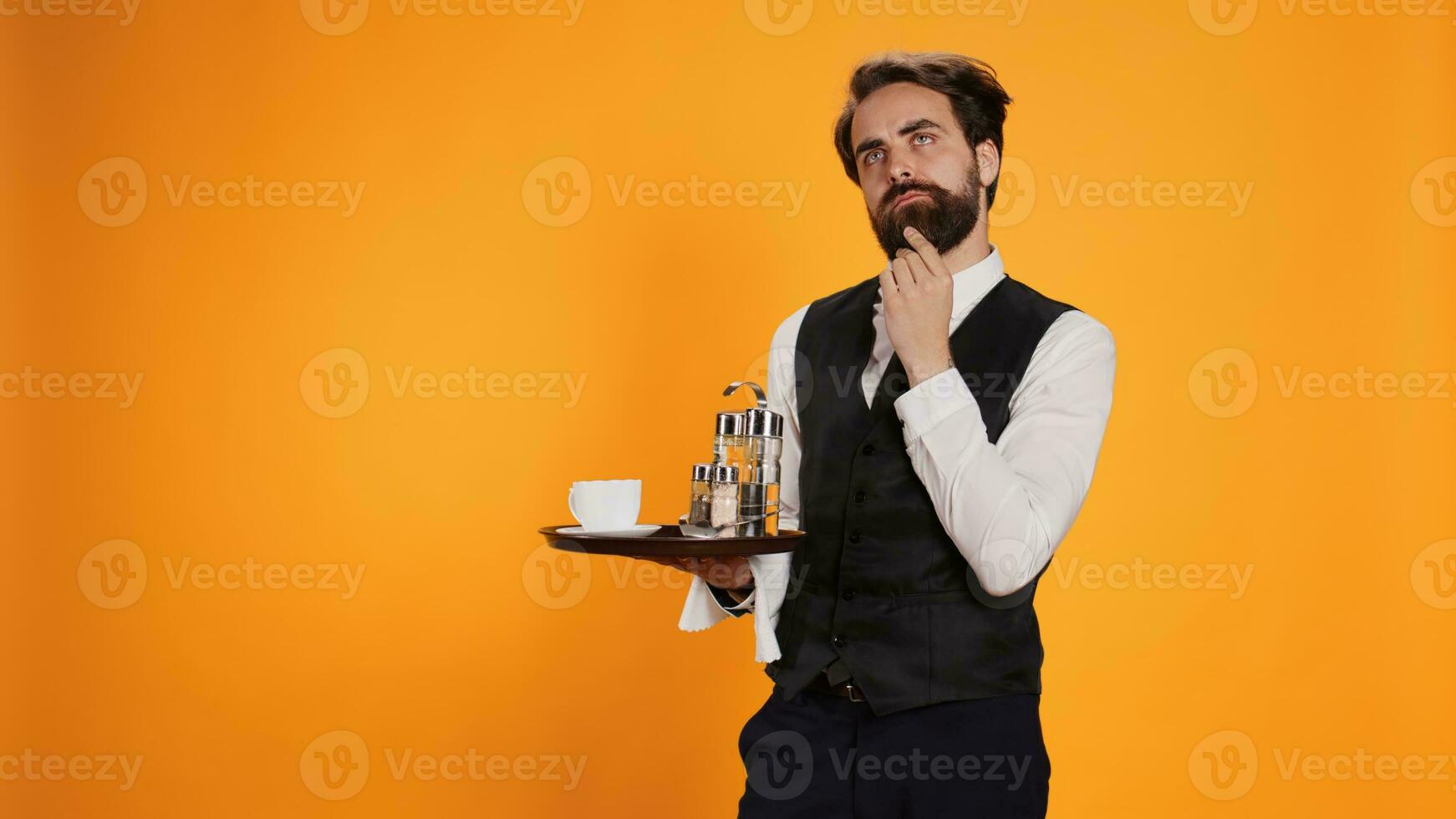 Thoughtful employee calculating check on camera, preparing to provide excellent service with food tray against yellow background. Elegant restaurant waiter being pensive on camera. photo