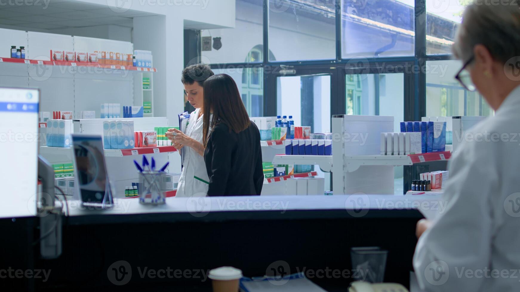 Asian client in need of prescription medicaments informations in apothecary, requesting help from cashier who redirects her to druggist checking pharmaceutical products inventory photo