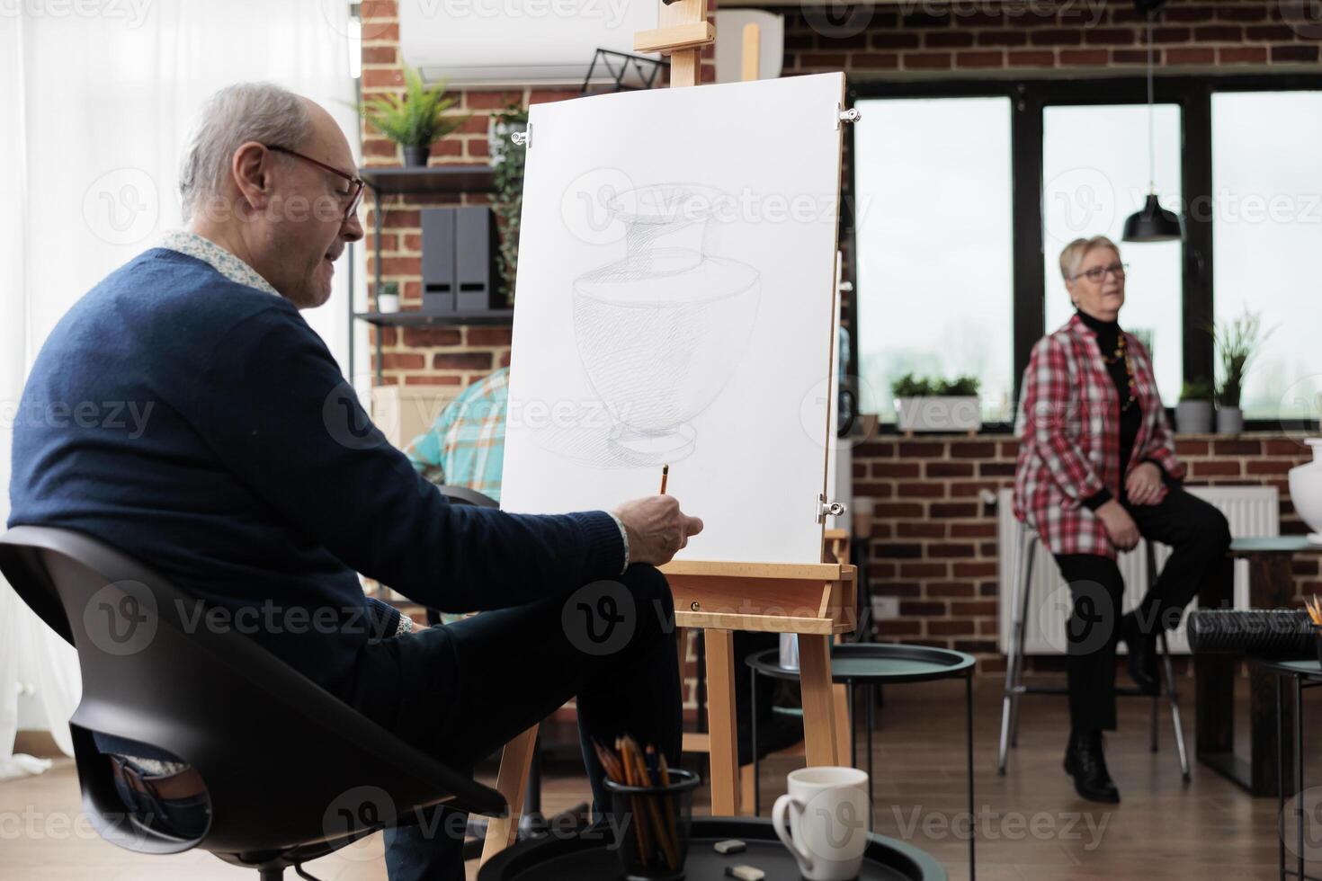 Senior man sitting at easel practicing drawing, visiting group art classes, learning new things in retirement. Mature male student sketching on canvas with pencil, attending creative arts activities photo