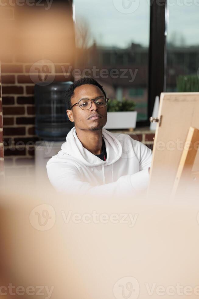 Inspired African American male student studying art at university, sitting at easel drawing from live model during workshop. Thoughtful black guy artist creating artwork, developing artistic skills photo