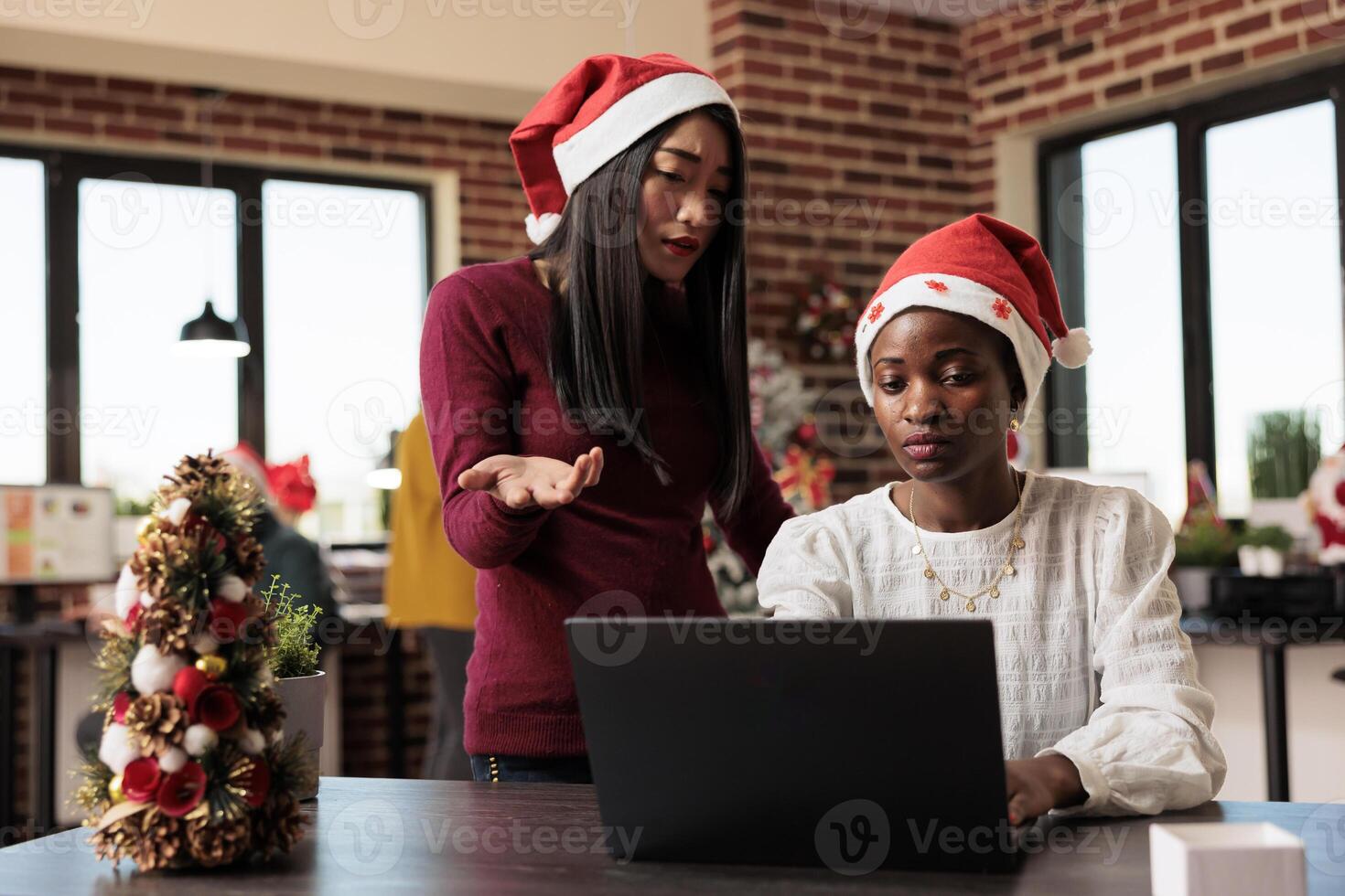 Asian and african american women working together on laptop in christmas decorated office. Project manager in santa hat helping colleague while using computer at festive workplace photo