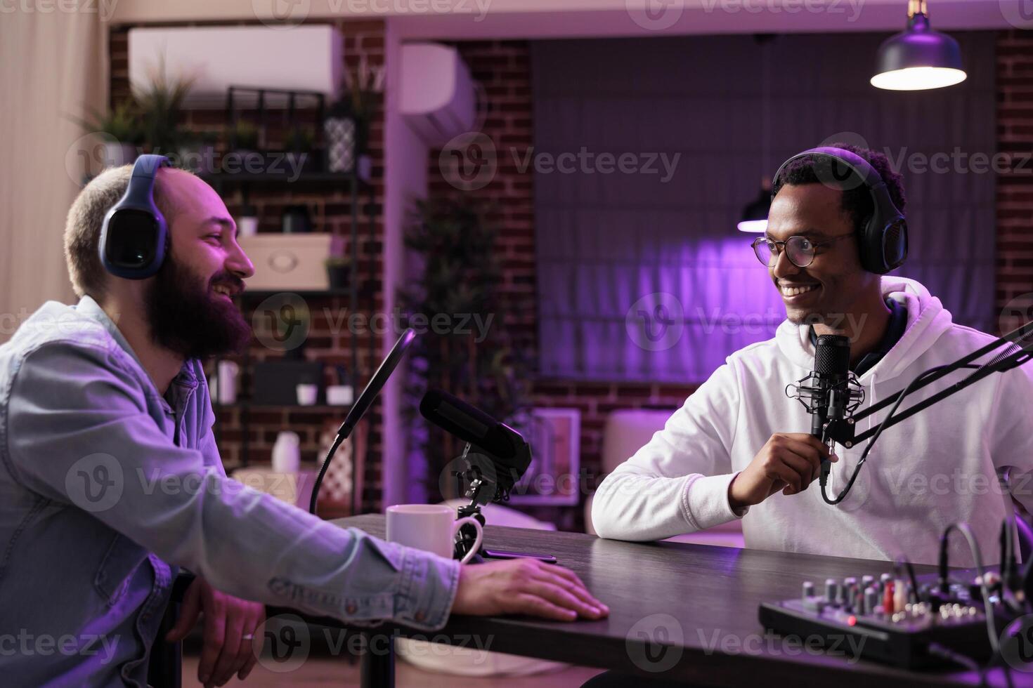Online show host recording podcast with guest, using audio recording equipment in home studio living room. Happy men laughing together during broadcasting session for internet streaming service photo