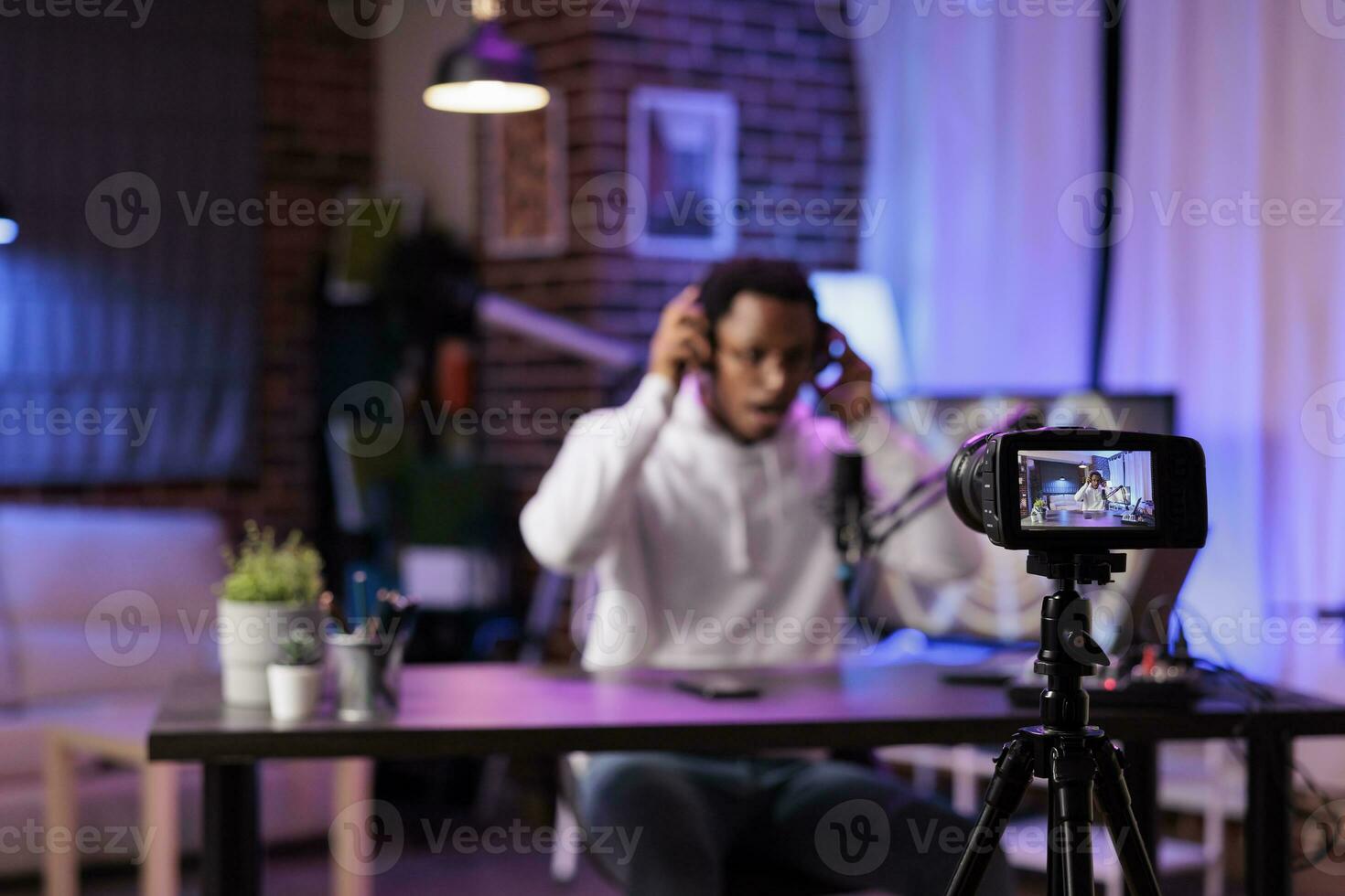 Focus shot on modern camera recording vlogger in blurry background talking about international news and events subjects. Man filming internet show using professional gadgets in rgb lights studio photo