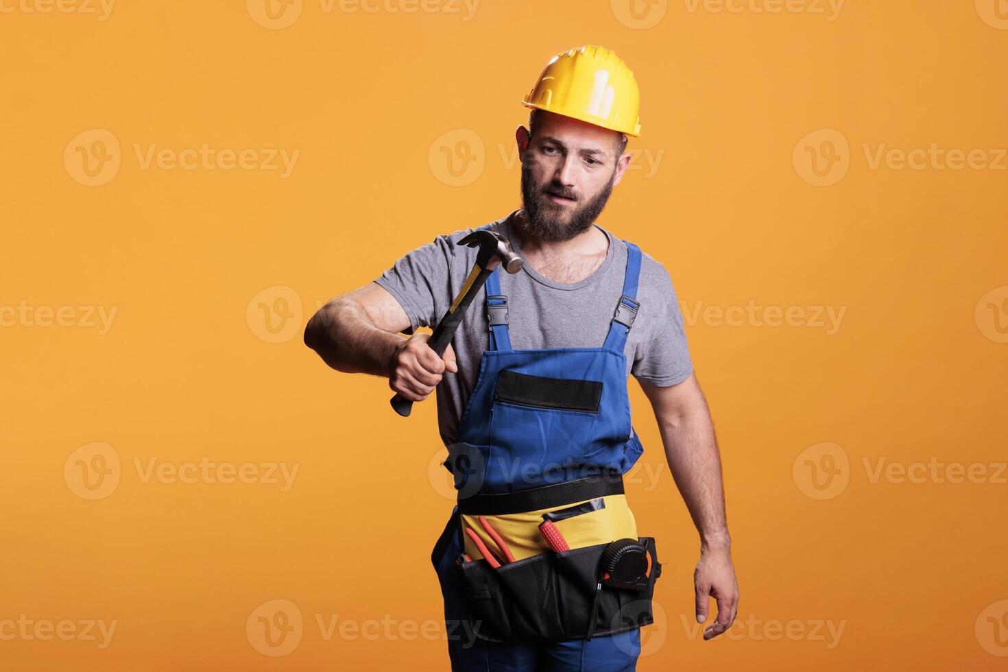 Construction worker using hammer on renovation project, standing over yellow background. Professional male renovator doing reconstruction work with sledgehammer and tools, studio shot. photo