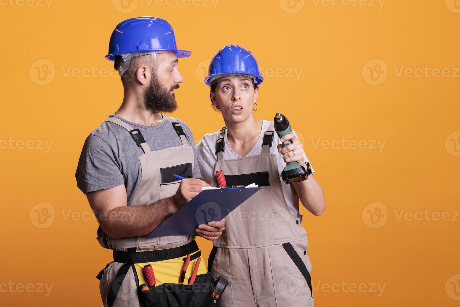 Team of construction workers taking notes on clipboard and checking renovation measurement, holding drill gun. Man and woman renovators looking at papers or files, standing over yellow background. photo