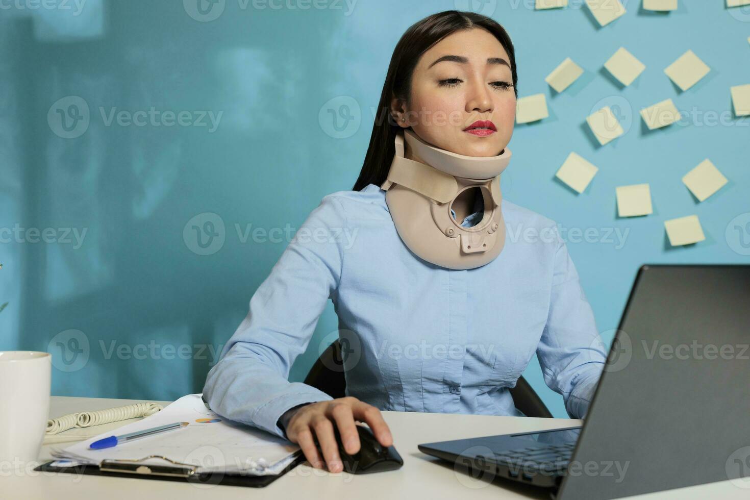 Corporate employee wearing medical collar for injury while working on reports, preparing paperwork for next meeting. Injured female entrepreneur concentrating on finishing project at work station. photo