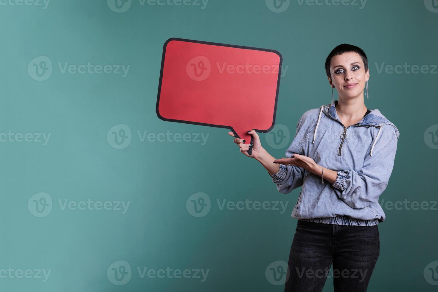 Cheerful model pointing at empty red speech bubble in studio, advertising copy space accessory for text message. Smiling attractive woman looking at camera while showing blank template board photo