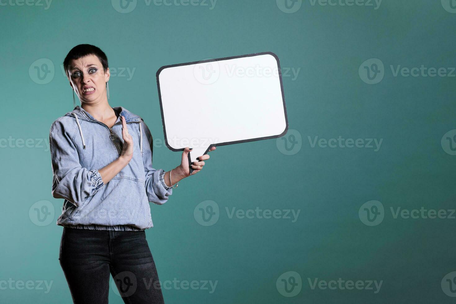 Cheerful brunette woman pointing at empty white speech bubble during studio shot, advertising copy space accessory for text message. Smiling model looking at camera while showing blank template board photo