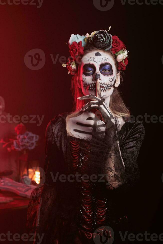 Spooky horror model doing hush sign to keep secret, wearing santa muerte halloween costume and showing slience mute and secrecy gesture with finger over lips, holy dios de los muertos. photo