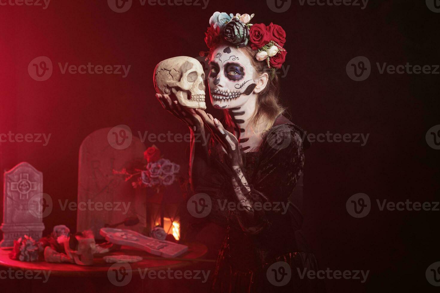 Santa muerte model holding skull in studio, acting scary and horror to celebrate mexican halloween. Beautiful woman in festival costume with body art, looking like goddess of death. photo