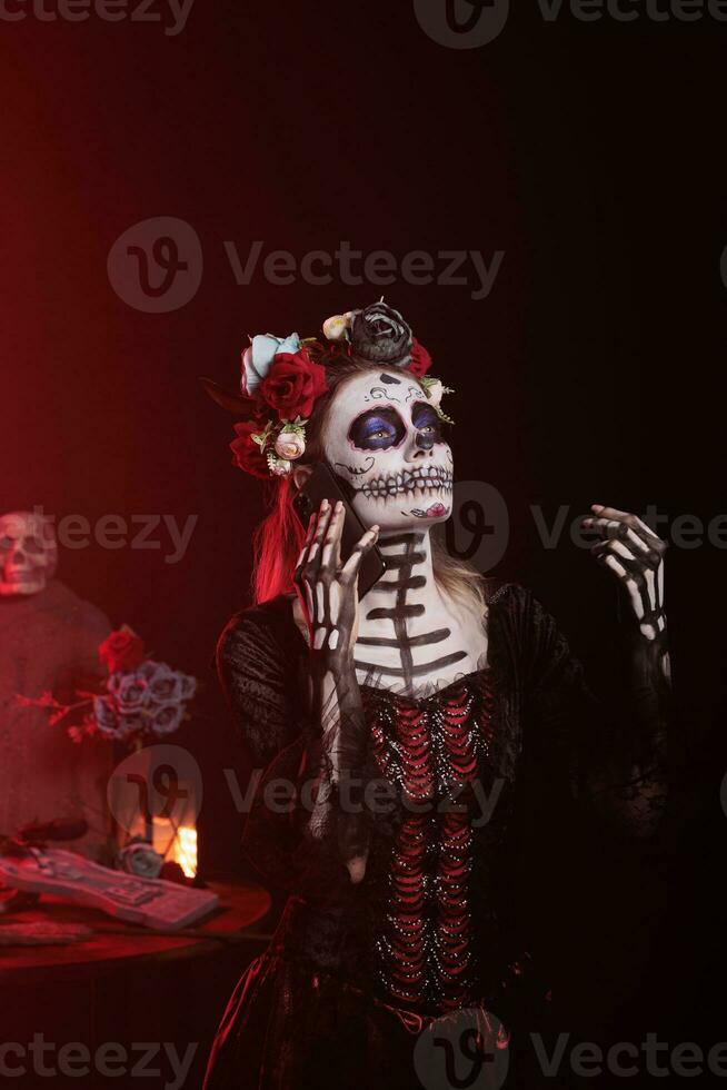 Goddess of death with sugar skull make up talking on phone call, using smartphone to chat in studio. Woman celebrating dios de los muertos holiday with la cavalera catrina costume, holy festival. photo